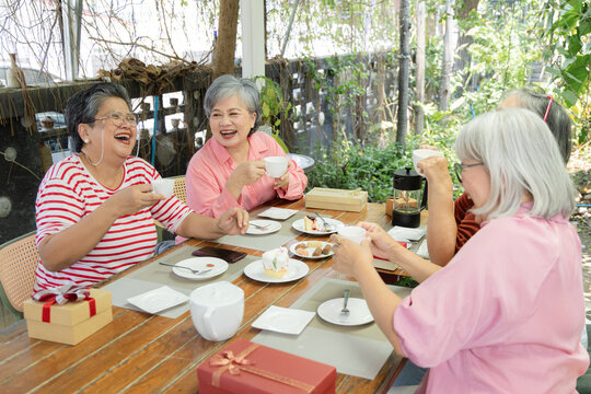 Active seniors laugh together over tea at garden cafe reunion, holding cups beside desserts, gift boxes while warm conversation and friendship create joyful outdoor social connection, aging society.