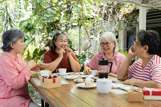 Senior women laugh together at garden cafe reunion, sharing tea, desserts, and friendly stories around table as wrapped gifts add cheerful celebration to relaxed outdoor gathering, aging society.