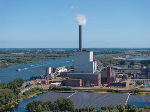 Aerial view of a large industrial power plant with a tall chimney emitting smoke, situated along a river with a floating solar panel array in the Netherlands.