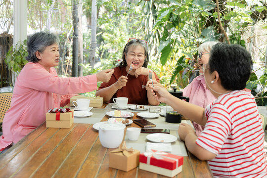 Active seniors share bites of cake during outdoor cafe reunion, smiling and laughing across table as desserts, tea, and gift boxes turn friendship gathering into joyful celebration, aging society.