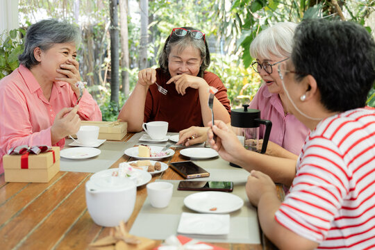 Retired senior women enjoy cake together at garden cafe reunion, laughing with forks in hand while desserts, tea, and presents create playful friendship and relaxed social connection, aging society.