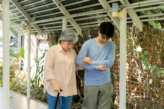 Elderly woman walks carefully with cane while young man supports her arm outdoors, showing family care, mobility assistance, confidence, safe walking during active senior daily life, aging society.