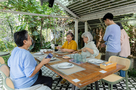 Retired senior women gather at outdoor cafe as young man assists nearby, while one friend uses smartphone and table is prepared for refreshments, conversation, relaxed social meeting, aging society.