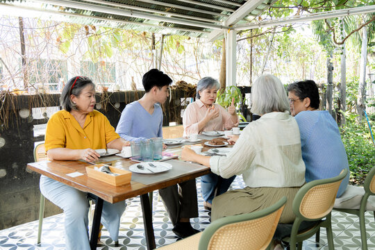 Senior women and young man enjoy casual garden cafe reunion, sharing snacks and conversation around table while relaxed gestures, friendship, and social connection shape warm gathering, aging society.