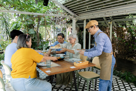 Elderly friends share plates as waiter serves food at outdoor cafe reunion, enjoying lunch, conversation around table while restaurant service supports relaxed friendship and community, aging society.