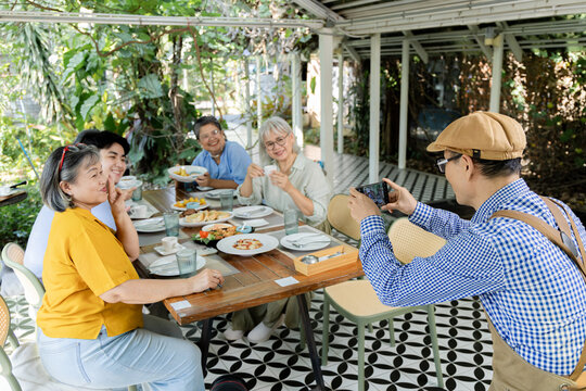 Waiter takes smartphone photo of senior friends at garden cafe reunion, capturing cheerful group with food and drinks while shared memories strengthen social connection outdoors warmly, aging society.
