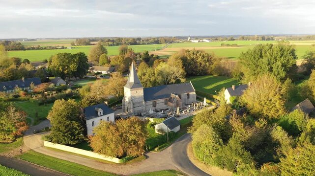 Aerial footage of the small rural town of Sasseville in Normandy, France, showing a traditional stone church with a steeple surrounded by houses, gardens, and lush green fields.