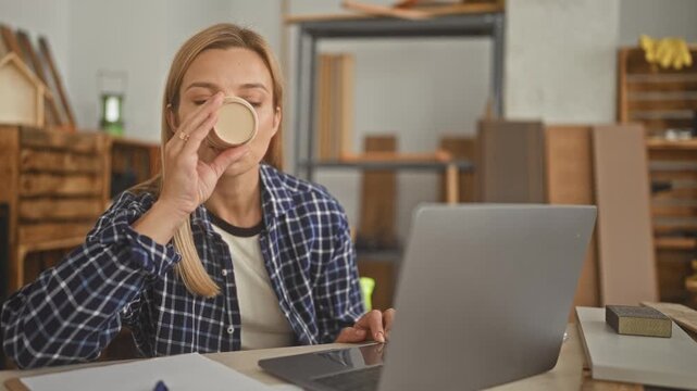 Woman drinking from paper cup at laptop with hand visible among wood panels and tools in a workshop building; focused work.