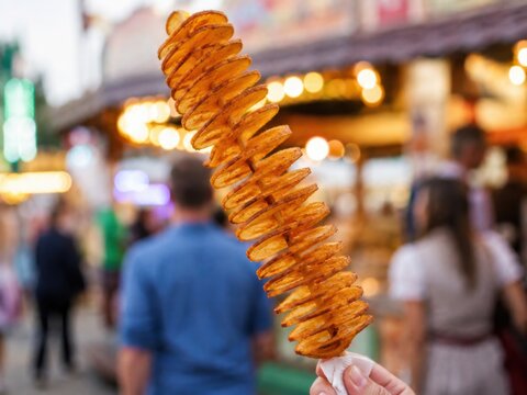 Knusprige Spiralkartoffel am Holzspie&szlig;, beliebter Snack auf Volksfest und Jahrmarkt. Goldbraun gew&uuml;rzt und vor einem belebten Verkaufsstand mit warmen Lichtern fotografiert.