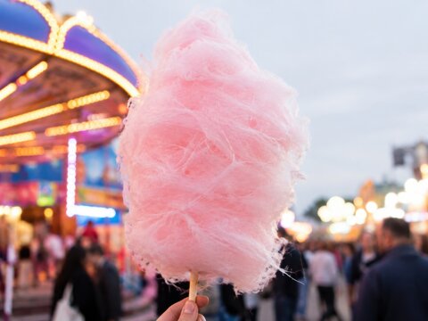 Rosa Zuckerwatte am Holzstab vor einem belebten deutschen Volksfest, s&uuml;&szlig;er Jahrmarktklassiker mit bunten Lichtern, Fahrgesch&auml;ft und unscharfen Besuchern in stimmungsvoller Atmosph&auml;re.