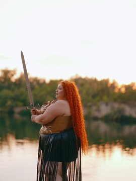 Side view of Plus Size Redhead Warrior Woman Holding Sword Outdoor by Lake