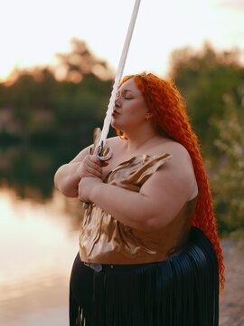Plus Size Redhead Warrior Woman Holding Sword Near Face by Lake with Closed Eyes