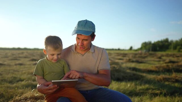 Father and son use tablet in field together. Happy boy touches tablet screen while farmer points. Father and child sit on hay bale outdoors. They explore digital tablet in rural farmland under sky.