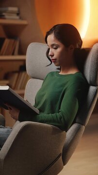 Young Asian woman reads scientific book in armchair, relaxing in home study