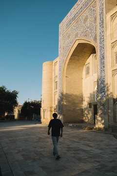 Man walking past a historic madrasah with blue tilework in Bukhara.