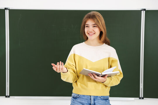 Teenage girl with book near chalkboard in classroom. Space for text