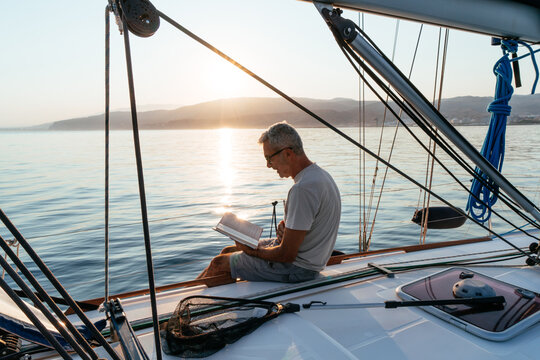 Senior man reading book on sailboat at sunset