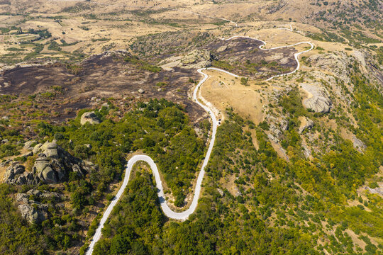 Aerial view of a winding asphalt road cutting through a hilly landscape with rocky outcrops, patches of scorched earth, and green vegetation in Dabnitsa, Prilep Municipality, North Macedonia.