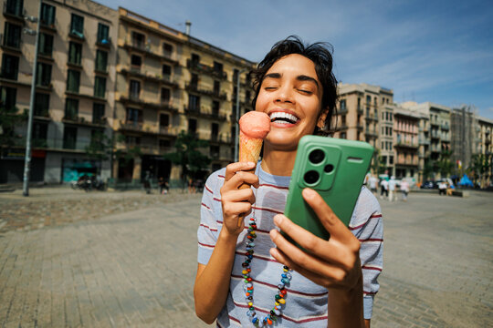 Woman enjoying ice cream and smiling while using smartphone