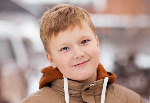 Young boy with light brown hair wearing a tan winter jacket smiles at the camera outdoors in a snowy environment with blurred background elements