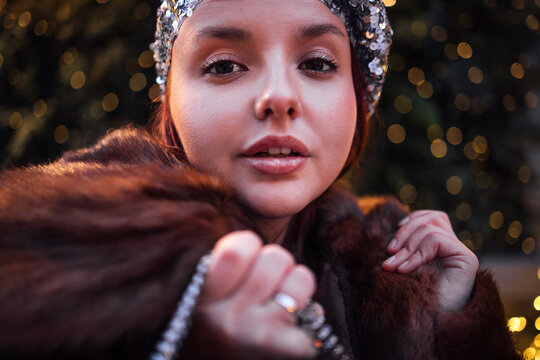 Young woman with sequined headband and fur coat poses for a close-up portrait against a backdrop of soft, blurred lights in an outdoor setting