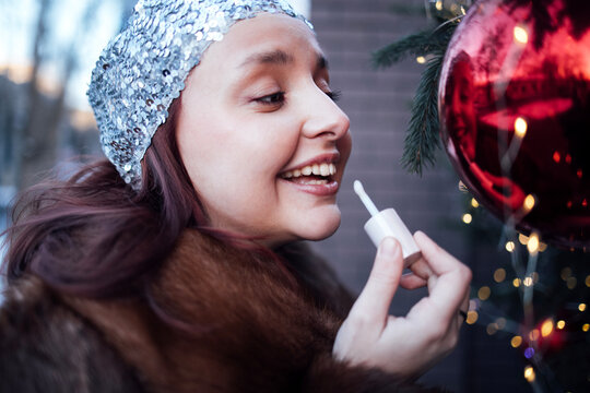 Young woman with silver headband applying lip gloss while smiling near a decorated Christmas tree with red ornaments and fairy lights in the background