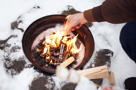 Person adding wood to a fire pit surrounded by snow, with flames visible and a skewer in hand for roasting marshmallows outdoors in a winter setting
