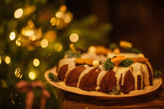 Festive bundt cake decorated with icing, orange slices, and pine branches on a wooden table, with a Christmas tree and warm bokeh lights in the background