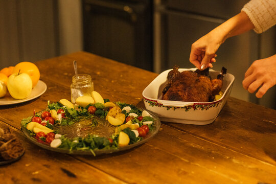 Woman's hand garnishes roasted chicken in a white serving dish on a wooden table, surrounded by a salad platter and fresh fruits in a cozy kitchen setting