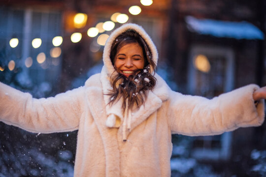 Young woman with long hair wearing a white fur coat smiles joyfully while standing in falling snow, illuminated by warm lights in a winter outdoor setting