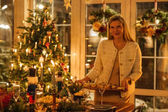 Woman in white cardigan holding a tray of festive treats in a decorated dining room with a Christmas tree and candles illuminating the warm atmosphere
