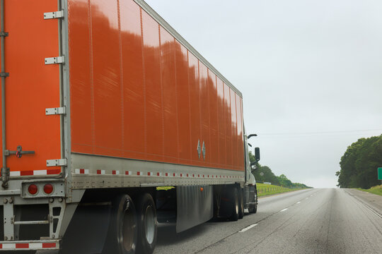 Large orange truck travels down highway with green trees on either side, under cloudy sky.