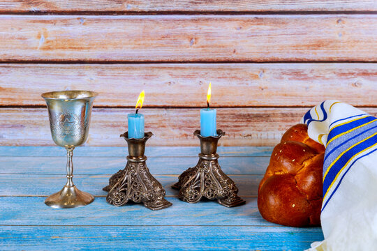 Ritual items are set on blue table two Shabbat candles, are lit beside cup, challah bread.