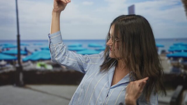 Woman mimes playing air guitar on a seaside street by blue beach umbrellas and the ocean, hair flying and hand strumming; joy celebration.