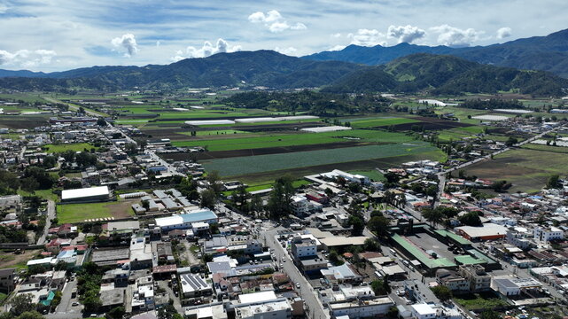 Aerial view of the Portezuelo area featuring agricultural fields, greenhouses, and mountain ranges under a blue sky in Constanza, La Vega, Dominican Republic.
