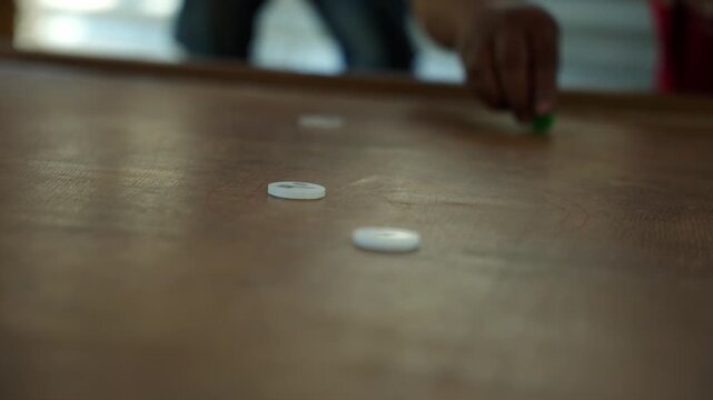Wide angle of scattered carrom pieces on worn wooden board with young man in red shirt leaning in during casual afternoon game session