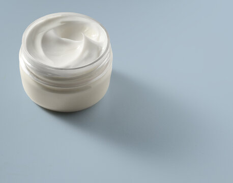 Open jar of white facial cream on pale blue surface with soft side lighting and minimal shadow