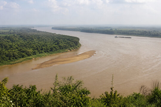 View from high bluff of wide brown river bending through low green forest, sandbar, tugboat and barge downstream, Mississippi River landscape