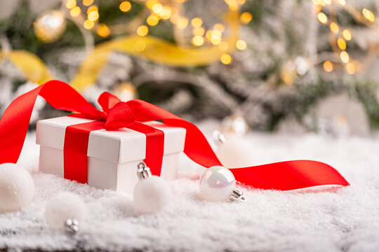 Festive white gift box wrapped with a red ribbon, resting on snow with pine branches, red baubles, and glowing holiday lights in the background. Ideal for holiday greeting cards, festive banners.