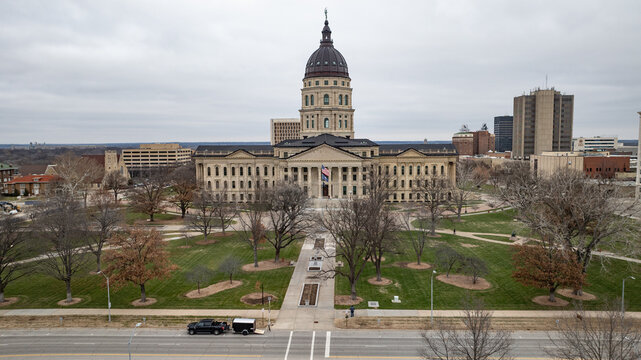 Aerial view of Kansas State Capitol surrounded by green lawns, bare trees, and city buildings under a cloudy sky in Topeka, Kansas, United States.