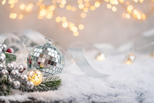 Reflective silver disco ball ornament rests on snow with golden ribbon, pine branches, and festive lights in a bright and cheerful holiday scene. Ideal for holiday greeting cards,