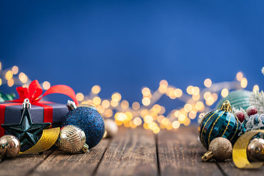 A Christmas background featuring navy and teal ornaments wrapped with gold ribbon on rustic wood, framed symmetrically against a deep blue backdrop with twinkling lights and copy space.