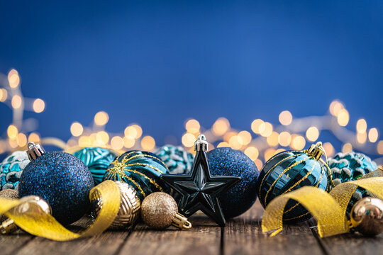 A Christmas background featuring navy and teal ornaments wrapped with gold ribbon on rustic wood, framed symmetrically against a deep blue backdrop with twinkling lights and copy space.