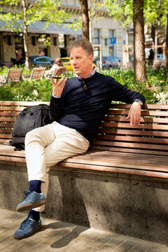 A middle-aged man is sitting on a bench in a park in a European city, using his phone