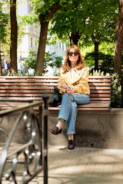 A middle-aged blonde woman sitting on a bench in a park in a European city
