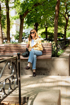 A middle-aged blonde woman sitting on a bench in a park in a European city