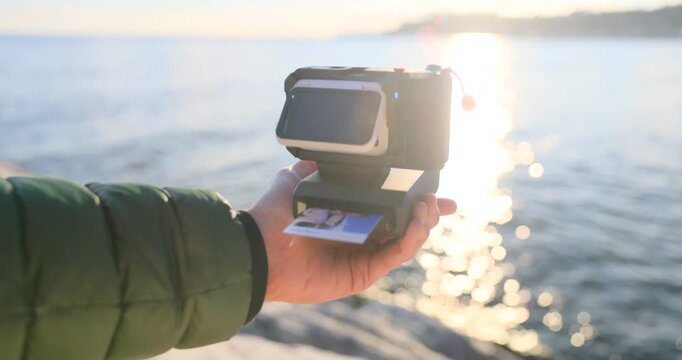 Hand holds an action camera with instant photo printer by the sea at sunset, printing a small snapshot. Creative travel gadget, vlogging gear and nostalgic memories concept