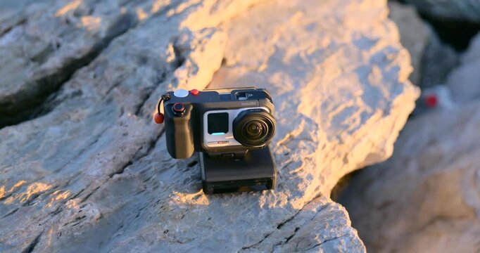 Orbit shot of a compact action camera mounted on sunlit coastal rocks at golden hour, highlighting rugged video gear