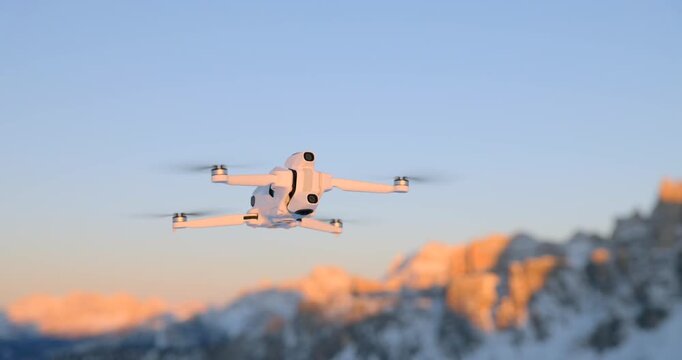 White quadcopter drone hovering in mid-air against snow-capped mountain peaks at golden hour sunset