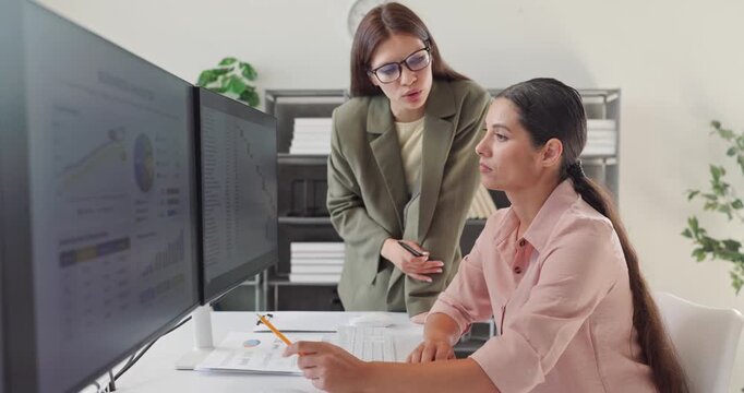 Women analysts discuss financial data analysis at computer. They review charts and KPIs on dual monitors as one colleague guides another. Image conveys leadership coaching and data teamwork.